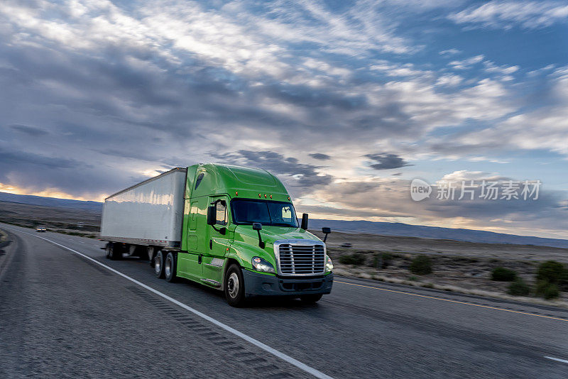 一辆大型商业货运半挂车在沙漠公路上向东行驶，以交付货物，一个戏剧性的日落背景