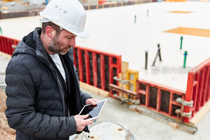 一名建筑师或土木工程师手拿iPad或平板电脑站在建筑工地上一个正在施工的地基前，头戴白色安全帽，身穿黑色夹克和羽绒服