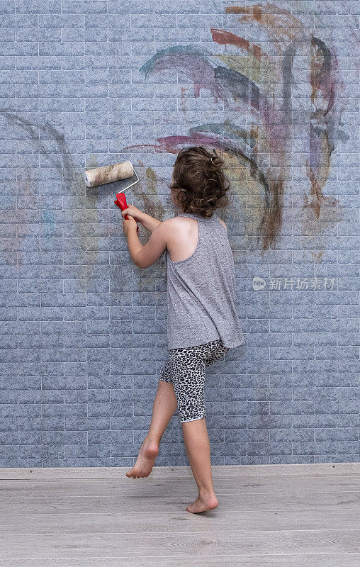 一个六岁的白人小女孩用油漆滚筒在墙上画画。装修房间。