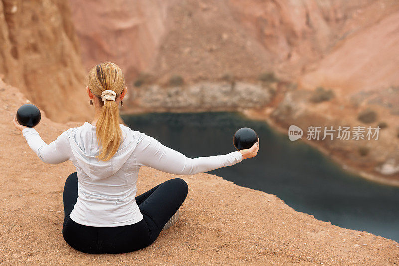 一名女子在狭窄的岩石之间的山湖前，抱着运动球，摆出瑜伽姿势