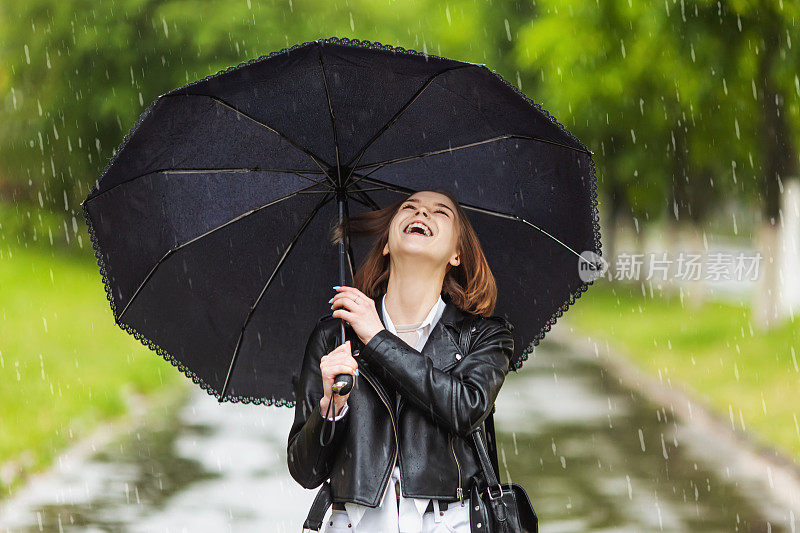 快乐的女孩穿着黑色夹克在春雨中漫步。在雨天打黑伞。下雨的城市。大雨在夏天的绿色背景。寒冷的秋天的天气。美丽的微笑