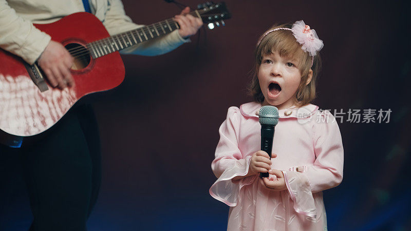 穿着复古礼服的小女孩在舞台上唱歌，她的父亲弹着原声吉他