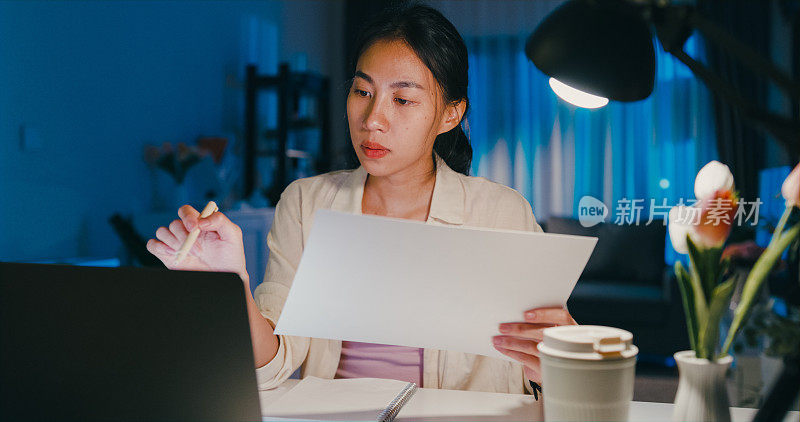 年轻的亚洲女性穿着正装衬衫坐在桌子前与笔记本电脑专注于屏幕和检查文件报告会议加班到午夜在家。