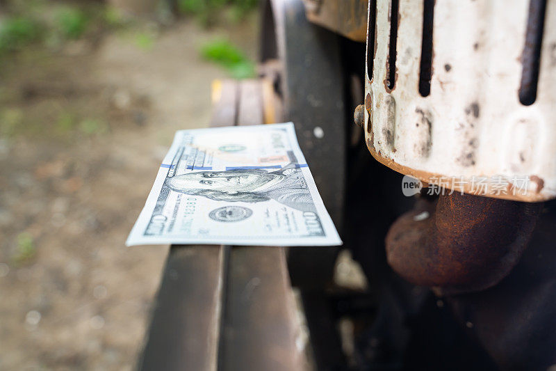 一张一百美元的钞票躺在一辆农村拖式拖拉机的发动机传动带上。钱在买旧柴油发动机的路上。农业机械美元