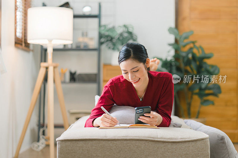 一位美丽的女商人躺在客厅的沙发上，一边工作一边用智能手机打电话，写笔记。
