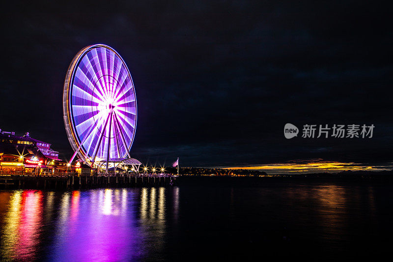 西雅图华盛顿西雅图摩天轮与复制空间在夜间被点亮，反映在太平洋港口69号码头长曝光灯在远处发光的阴天金色云