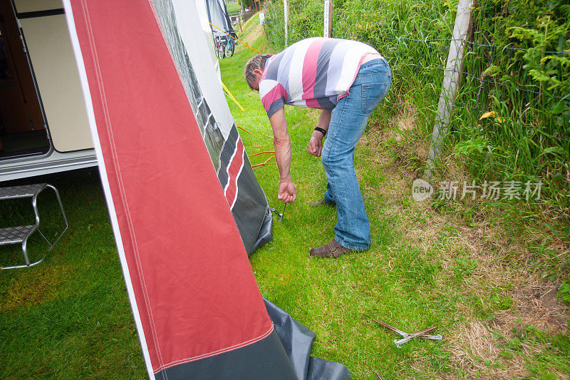 夏日，一名男子在威尔士乡村用钉子固定遮阳篷，准备在大篷车和遮阳篷中度假，为家人提供更多的使用空间。