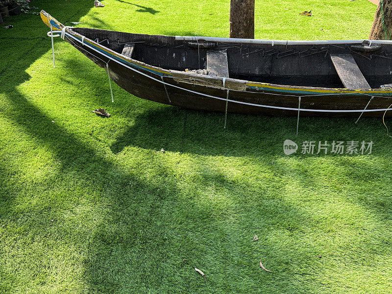 图片上的木制渔船船头拖在人造的绿色草坪上，阴影投射在阳光下，聚焦于前景