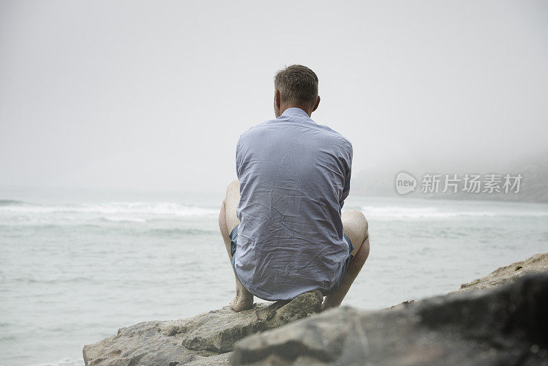 一个男人坐在岩石上，眺望着雾蒙蒙的海滩