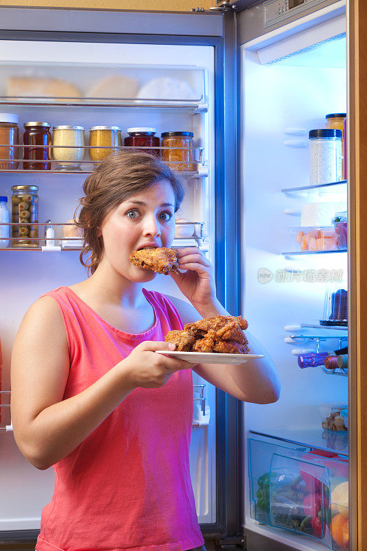 一个女人在打开的冰箱前吃不健康的炸鸡