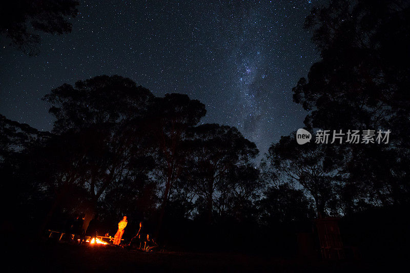 一群露营者围着篝火站着，上面闪烁着明亮的星星