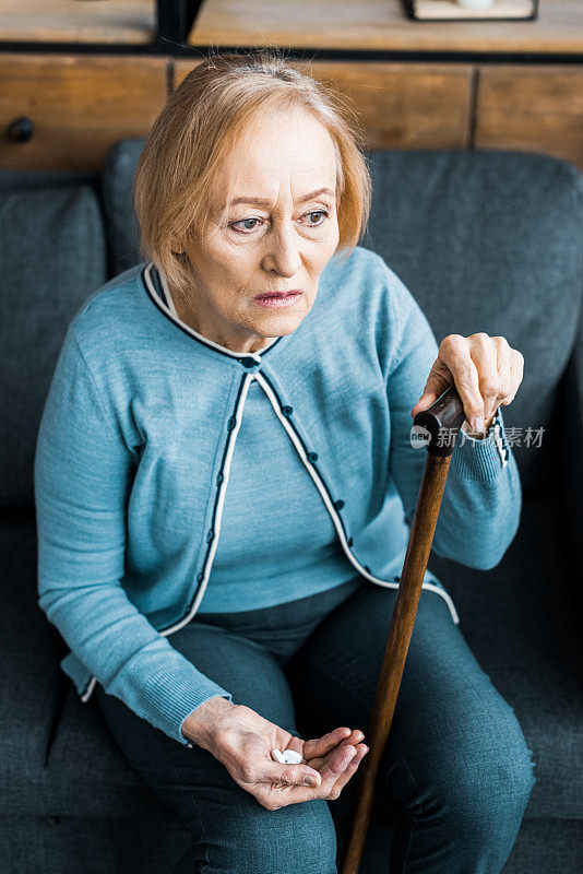 生病的老妇人拄着拐杖坐在沙发上，手里拿着药