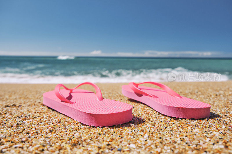 海滩假日背景，粉色人字拖在金色的沙滩上，映衬着蓝色的天空和大海，康沃尔，英格兰。