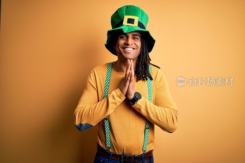 年轻的非洲裔美国人戴着绿帽子庆祝圣帕特里克节，双手一起祈祷，自信地微笑，请求宽恕。