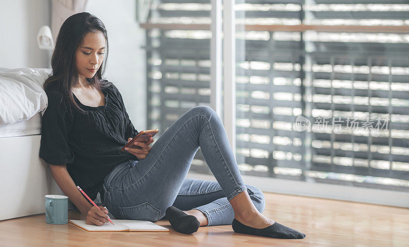 年轻迷人的亚洲女性穿着黑色衬衫和蓝色牛仔裤在家工作，坐在卧室的地板上使用智能手机和写作。