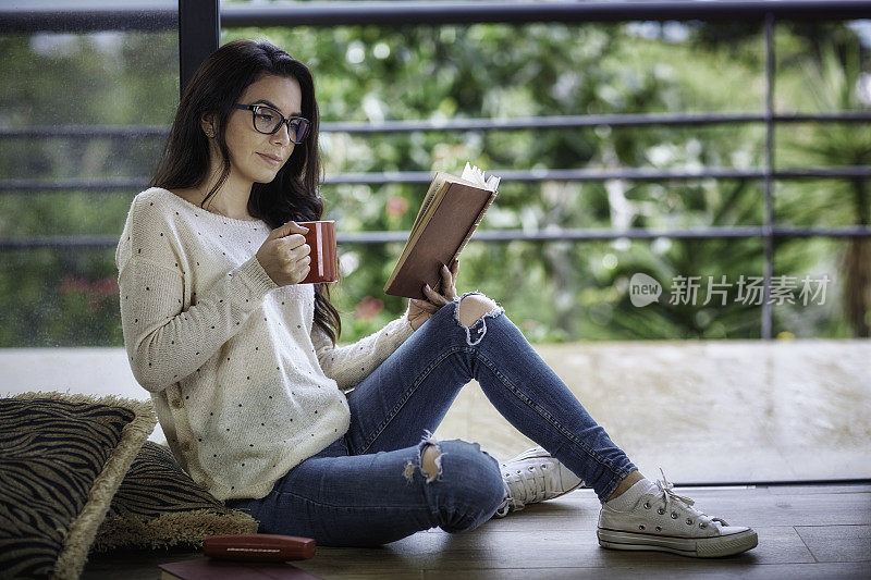 西班牙裔年轻女子戴着老花镜在家看书。坐在地板上。