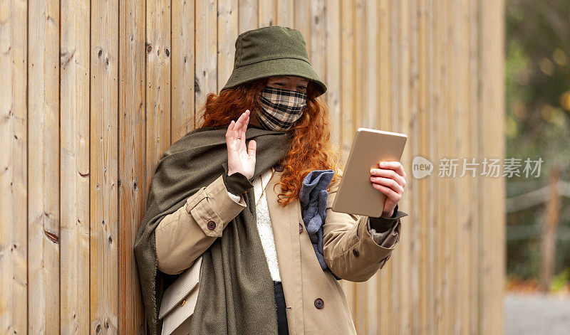 一名十几岁的女学生戴着一个保护面罩，在一个视频电话会议中向她的家人挥手，站在公园的一堵木墙旁边