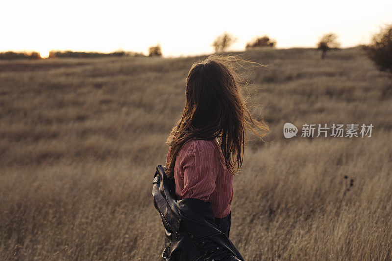 一位年轻女子在高高的草地上脱下她的皮夹克，欣赏日落