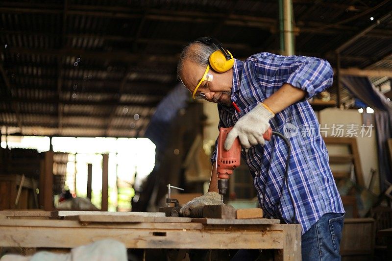 年长的亚洲木匠是用手钻与木板在他自己的车库风格的车间在退休年龄的爱好