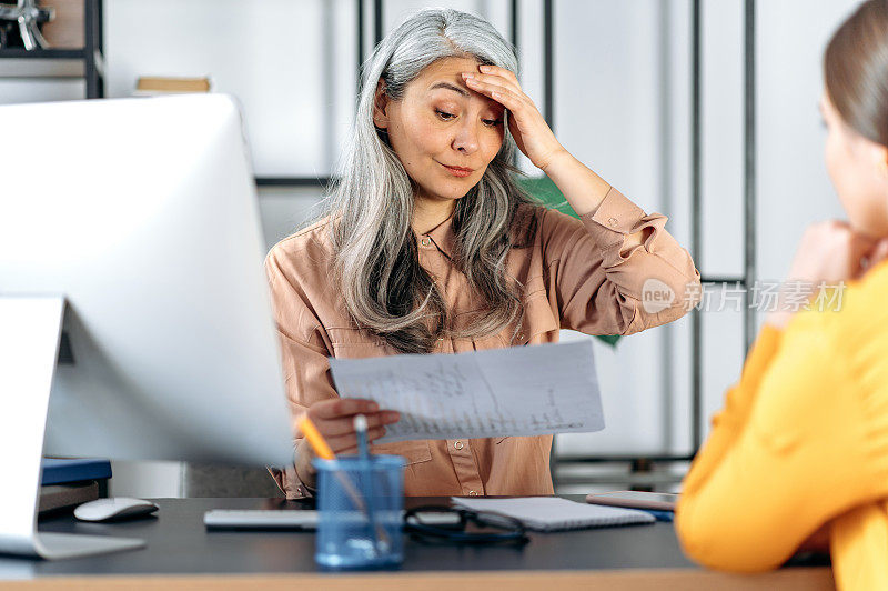一个恼怒气馁压抑成熟的灰白头发的亚洲女人，商业女人，女老板，在一个现代化的办公室，对工作不满，责骂一个雇员在财务报告中的错误