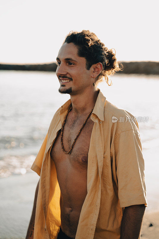 快乐英俊的嬉皮士意大利男子的肖像与长发微笑，在日落时走在海滩上。旅游热。