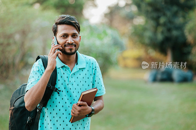 年轻的男学生在公园用书包和书使用智能手机