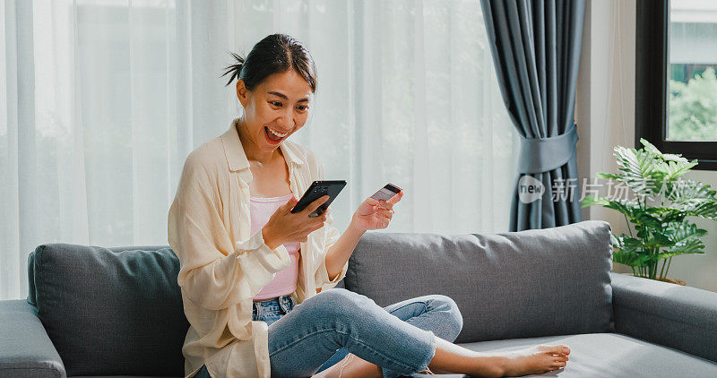 年轻的亚洲女女孩穿着休闲坐在沙发上，抱着智能手机和信用卡感觉兴奋和幸福的网上购物成功支付在客厅在家里。