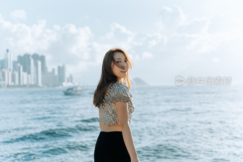 在香港海岸线的景色前，亚洲年轻女子回望镜头，微笑着