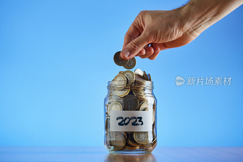 一个人的手拿着两枚硬币放在硬币储蓄罐里，标签上写着2023的文字，装满了硬币