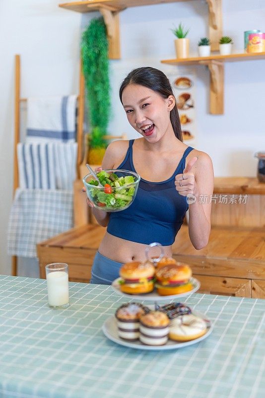 照片年轻美丽的亚洲女性女士在运动服装说不和拒绝诱惑汉堡和甜甜圈在厨房的桌子上，选择一碗沙拉与樱桃番茄和一杯牛奶的饮食和减肥健康的生活方式