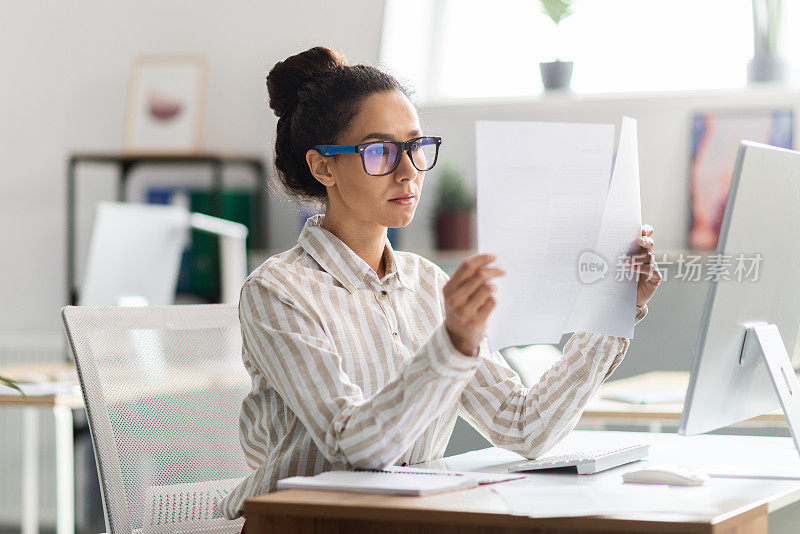 戴眼镜的严肃忙碌的女人坐在桌子前，用电脑在办公室内部处理文件