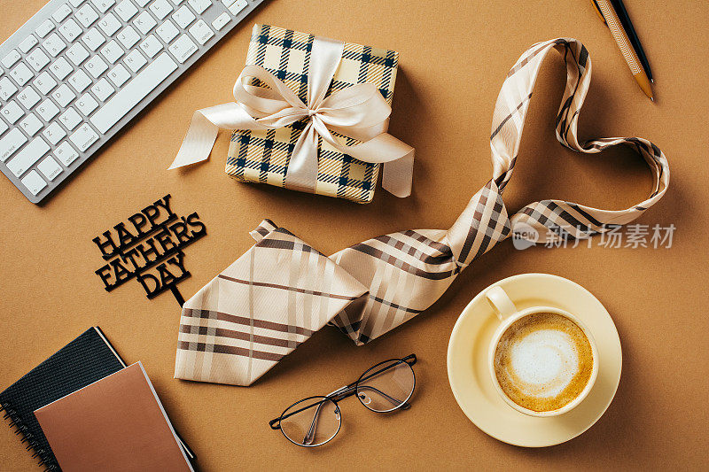 父亲节快乐的概念。平铺构图用心形领带、咖啡杯、礼盒、眼镜、键盘、棕色背景的纸质笔记本