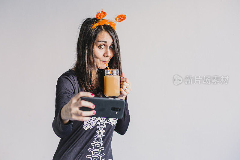 年轻美女喝着橙汁，拿着手机自拍。穿着黑白相间的骷髅服。万圣节的概念。在室内