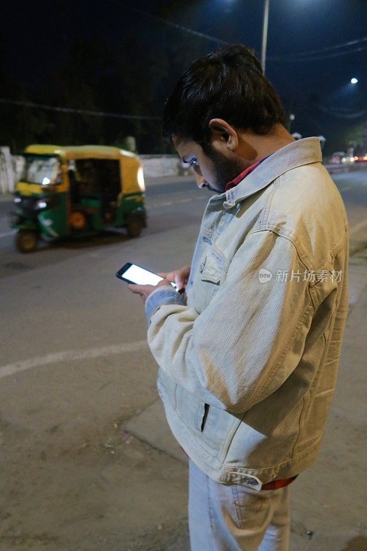 印度男子晚上用手机打电话，打车出租，双牛仔夹克牛仔裤，印度男子看手机屏幕由绿黄相间的机动三轮车，新德里嘟嘟的背景，交通公共交通印度