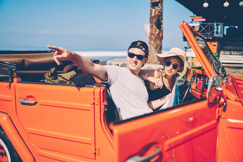 年轻男子在太阳眼镜指在公路上坐在租来的汽车和女朋友一起娱乐，时髦的夫妇探索热带岛屿的cabriolet有公路冒险在夏天