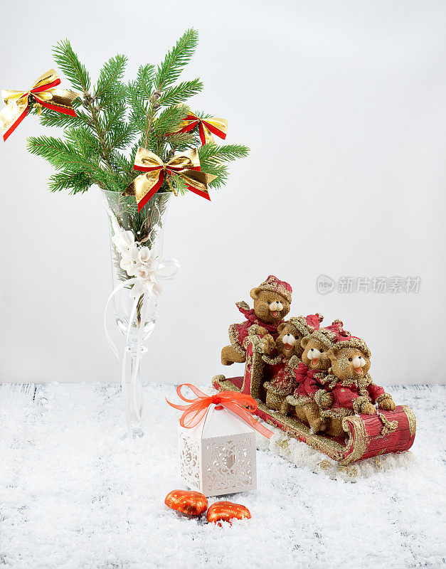圣诞树上装饰着白色的高脚杯，白色的礼盒，红色和金色的玩具熊在雪橇上，糖果和雪在复古复古的白色桌子上，白色的背景