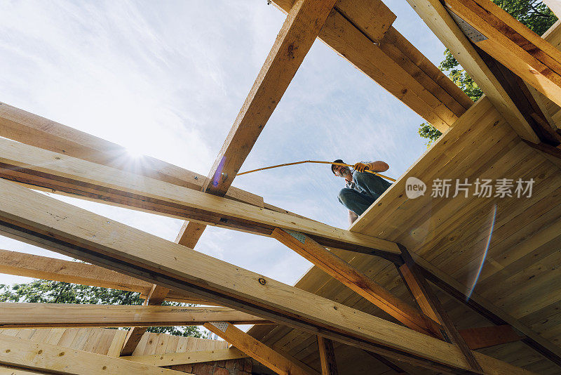 在施工现场，面对天空，成熟的男木匠手持卷尺站在木屋梁上