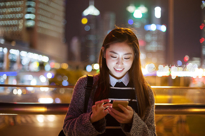 年轻美丽的亚洲女人晚上在现代城市玩手机