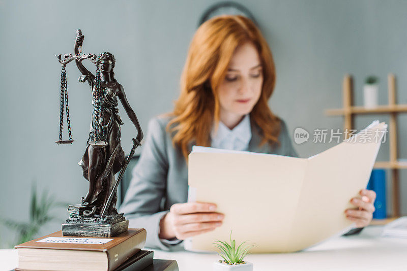 女律师看文件夹，而坐在工作场所与themis雕像在一堆书模糊的背景