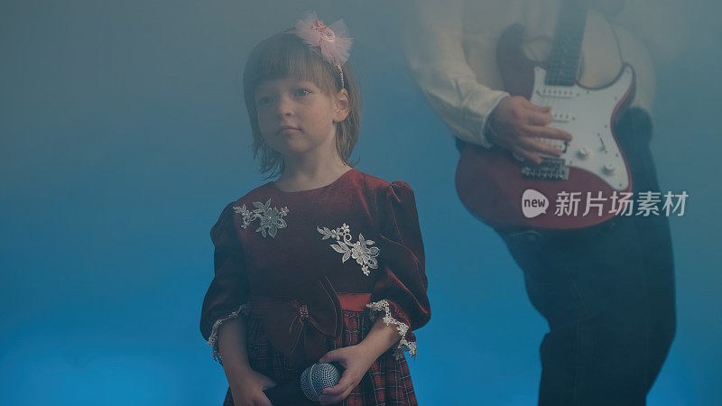 穿着复古礼服的小女孩在舞台上唱歌，她的父亲在弹奏电吉他