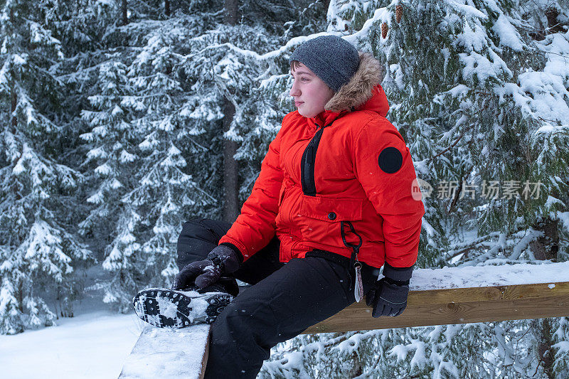 以白雪皑皑的森林为背景，一名身穿冬季红色夹克的白人青年望向远方。