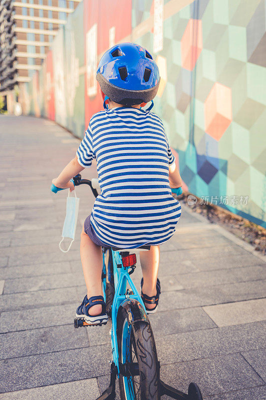 在新冠肺炎疫情期间，可爱的小男孩戴着防护口罩和安全帽在阳光明媚的夏日在公园骑自行车，新常态的生活方式概念。