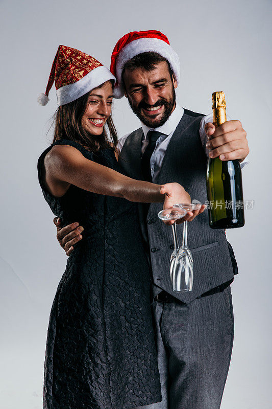 在节日的工作室背景下，一对穿着西装的夫妇在五彩纸屑和香槟的包围下戴着圣诞老人帽，为他们的庆祝活动增添了一丝异想天开。