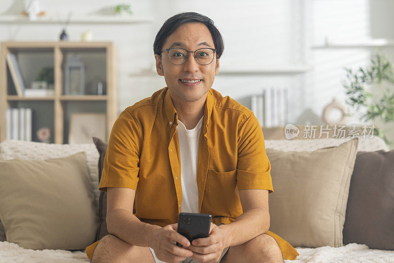亚洲男性智能手机短信聊天，亚洲成年青少年微笑着愉快地与朋友聊天，坐在沙发沙发客厅室内。