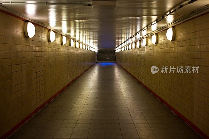 空荡荡的人行地铁隧道，黄色的灯光和瓷砖墙通向一个蓝色的出口标志。