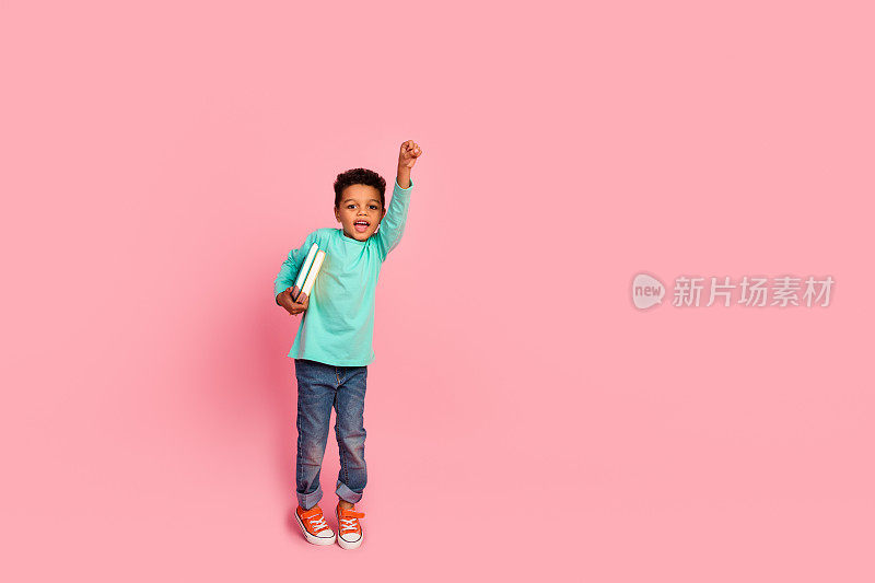 可爱的小男孩举起拳头，超级英雄拿着书，穿着时髦的海蓝宝石服装，在粉红色的背景上孤立起来