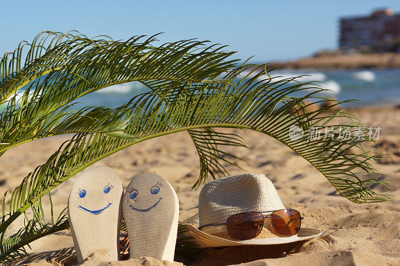 沙滩拖鞋上画着笑脸，帽子上戴着太阳镜，放在海边的棕榈叶下