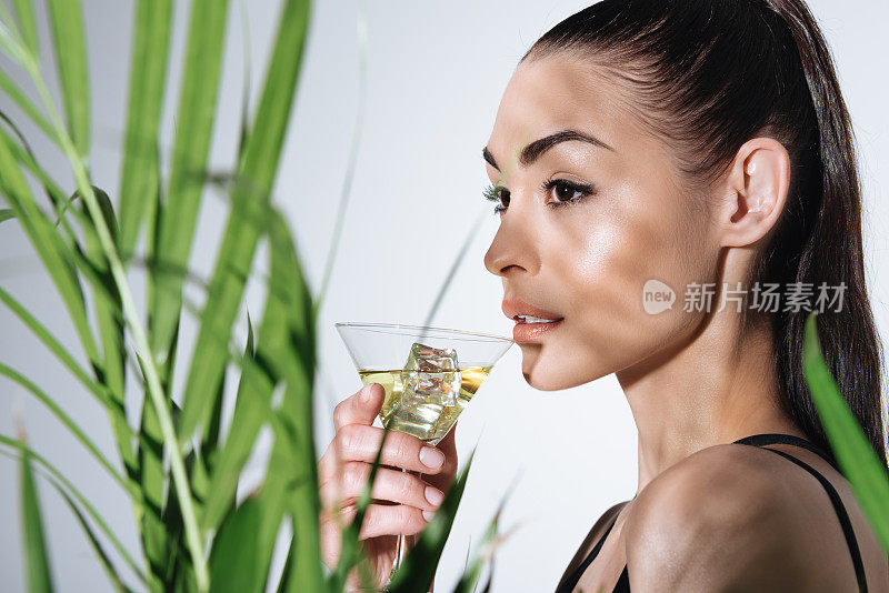 美丽年轻的黑发女人喝鸡尾酒和摆姿势附近的绿色棕榈叶