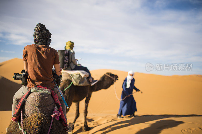 日落时分，游客骑着骆驼穿过撒哈拉沙漠的沙丘——摩洛哥。