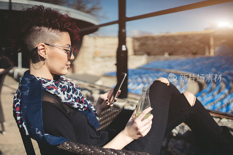 漂亮的红发女人坐在一个空的露天剧院享受最喜欢的饮料和使用手机。